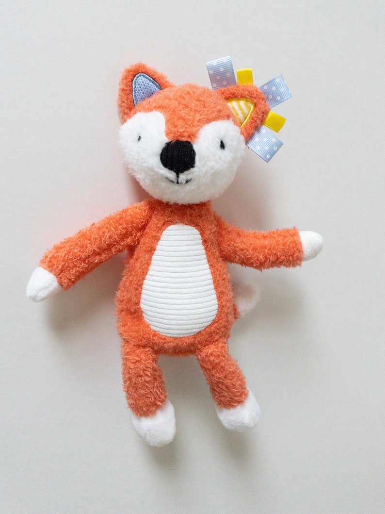 Puede incluir: Un peluche de zorro con pelaje naranja texturizado, cara blanca y vientre blanco con rayas horizontales. El zorro tiene una nariz negra, patas blancas y cintas azules y amarillas unidas a una oreja. Las orejas tienen detalles en azul y amarillo.