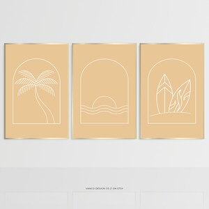 May include: Set of three minimalist line art prints with a beach theme. The prints feature a palm tree, a sunset over the ocean, and surfboards with palm leaves. The prints are framed in gold and have a light beige background.
