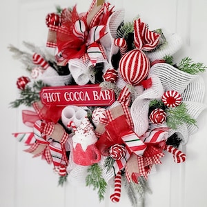 May include: A red and white Christmas wreath with a sign that says "Hot Cocoa Bar". The wreath is decorated with candy canes, red and white striped balls, and red bows.