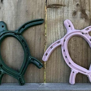 以下が含まれることがあります： 4つの蹄鉄アート作品。カメの形をしており、紫、緑、ピンクの色合いで塗装されています。背景は風化した木製のフェンスです。