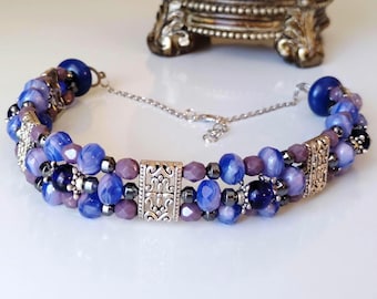 May include: A multi-strand bracelet featuring blue and purple marbled beads, small dark metallic spacer beads, and rectangular silver-toned metal filigree accents. The bracelet includes a silver-toned chain extender with a lobster claw clasp.