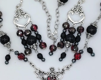 May include: A jewelry set consisting of a necklace and matching earrings. The necklace features a silver-tone metal pendant with a red heart and rose motif, accented with black and deep red beads. The earrings mirror this design with silver heart-shaped connectors, black beads, and faceted red crystals.