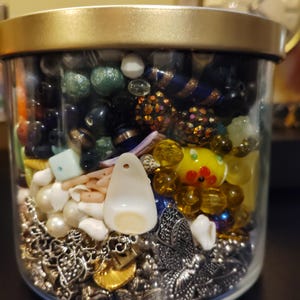 May include: A clear glass jar filled with a variety of beads, charms, and small decorative items. The contents include pearls, silver charms, seashells, and colorful glass beads in shades of blue, green, yellow, and black. The jar has a gold-colored lid.
