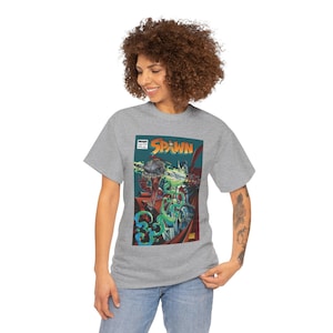 Peut inclure: Un t-shirt gris chiné à manches courtes avec une impression graphique représentant une couverture de bande dessinée Spawn. L'illustration montre un personnage sombre et armuré avec une cape rouge flottante et un effet d'énergie verte.