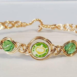 Op de afbeelding: Goudkleurige draadarmband met een geweven ontwerp. De armband heeft een centrale cirkelvormige bedel met een bloemenontwerp en twee groene kristallen kralen. De sluiting is een eenvoudige haak- en oogsluiting.