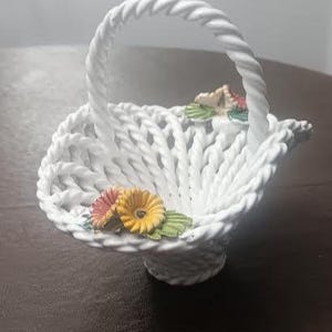 May include: A white ceramic basket with a woven design and a twisted handle. The basket is decorated with colorful ceramic flowers, including yellow, pink, and green accents. The basket is approximately 15 cm in diameter.