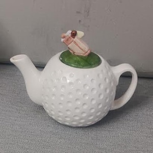 Op de afbeelding: Witte keramische theepot met een golfbaltextuur. Het deksel heeft een groen blad en een miniatuur golftas. De theepot heeft een gebogen tuit en een afgeronde handgreep. Een uniek cadeau.