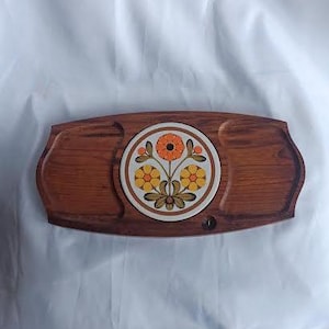 May include: A vintage wooden serving tray with a central circular tile featuring a floral design in orange, yellow, and brown. The tray has a rectangular shape with curved edges and two recessed areas. A small hole is visible on the right side.