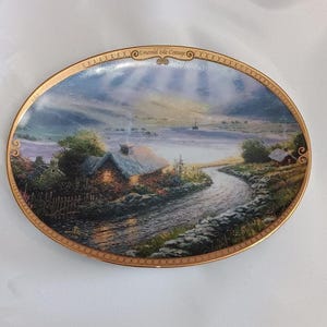 May include: Oval decorative plate featuring a scenic landscape with a cottage, river, and sunset. The plate has a gold rim and the text "Emerald Isle Cottage". The artwork depicts a serene, rural scene with soft colors.