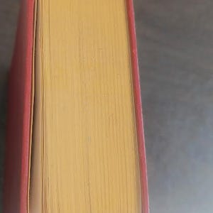 May include: A vintage hardcover book with a red cover and yellowed pages. The book's pages are visible, showing the texture and age of the paper. The book is likely antique.