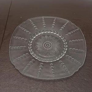 May include: Clear glass plate with a decorative pattern. The plate features a central sunburst design surrounded by a beaded border and radiating beaded lines. The plate has a scalloped edge.