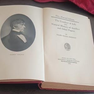 May include: An open book with a red spine. The left page features a black and white portrait of Thomas Carlyle in an oval frame. The right page displays the title "The Conduct of Life and Natural History of Intellect and Other Papers" by Ralph Waldo Emerson.