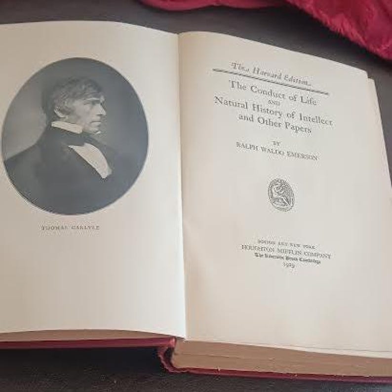 May include: An open book with a red spine. The left page features a black and white portrait of Thomas Carlyle in an oval frame. The right page displays the title "The Conduct of Life and Natural History of Intellect and Other Papers" by Ralph Waldo Emerson.