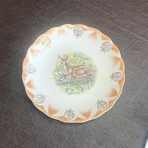 Puede incluir: Un plato de cerámica blanco con borde festoneado y una escena de ciervos en el centro. El borde tiene un ribete color melocotón con detalles florales decorativos. El plato es redondo y parece vintage.