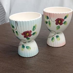 May include: Two ceramic egg cups with a floral design. The cups are white with a scalloped rim and a pedestal base. Each cup is decorated with a red flower and green leaves. One cup has a blue rim, and the other has a pink rim.
