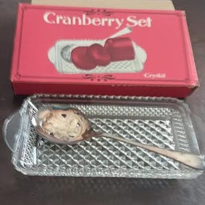 Puede incluir: Un juego de arándanos con una caja roja con la inscripción "Cranberry Set". El juego incluye una cuchara de servir plateada y una bandeja de cristal transparente, rectangular, con un patrón de diamantes.