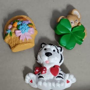 May include: Three small decorative figurines: a tan woven basket filled with colorful flowers and a pink bow, a brown mouse peeking from behind a large green four-leaf clover, and a white tiger with black stripes, a red bow, and a red heart.