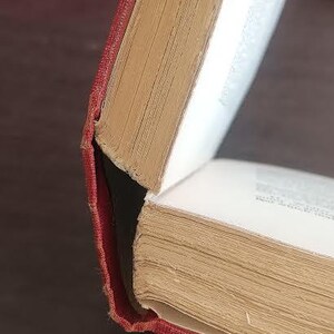 May include: Close-up of an open book with a red cover and aged, yellowed pages. The book's spine is visible, and the pages are slightly fanned out, revealing the text within. The book is held open.