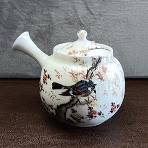 May include: A white ceramic teapot with a side handle and a matching lid. The body features a detailed illustration of a bird perched on a branch, surrounded by delicate red and brown floral patterns with subtle gold accents throughout the design.