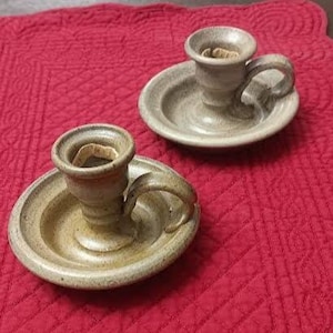 May include: Two handmade ceramic candle holders. Each candle holder has a circular base with a handle and a cup-shaped top. The candle holders are a speckled tan color and sit on a red quilted surface.