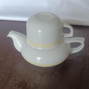 May include: A white ceramic tea set consisting of a teapot and a stacked cup. The teapot features a wide base with a handle and spout, while the cup sits securely on top. Both pieces are decorated with a thin, horizontal yellow band.
