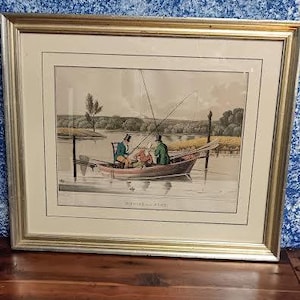 May include: Framed vintage print depicting a fishing scene with three people in a boat on a lake. The print is in a gold-colored frame with a white mat. The boat has fishing poles and is surrounded by water and trees.
