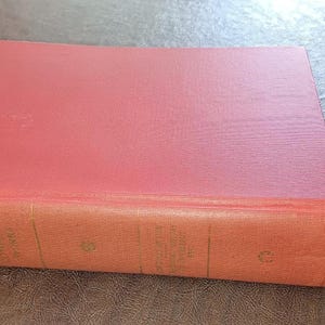 May include: A vintage book with a red cover and aged, orange-toned edges. The book is rectangular and appears to be hardcover. The cover has a slightly textured appearance, suggesting it may be made of cloth or a similar material. The book is resting on a brown surface.