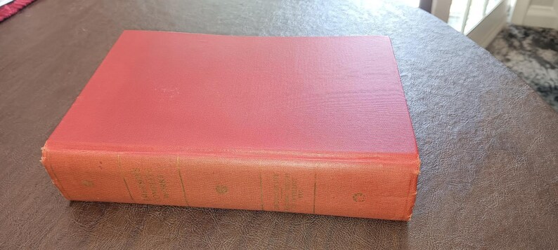 May include: A vintage book with a red cover and aged, orange-toned edges. The book is rectangular and appears to be hardcover. The cover has a slightly textured appearance, suggesting it may be made of cloth or a similar material. The book is resting on a brown surface.