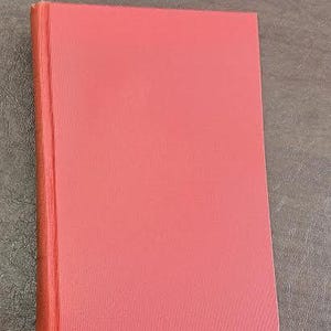 May include: A closed, hardback book with a solid coral-coloured cover. The spine is visible on the left side, and the pages are a similar shade. The book is rectangular and sits on a textured surface.