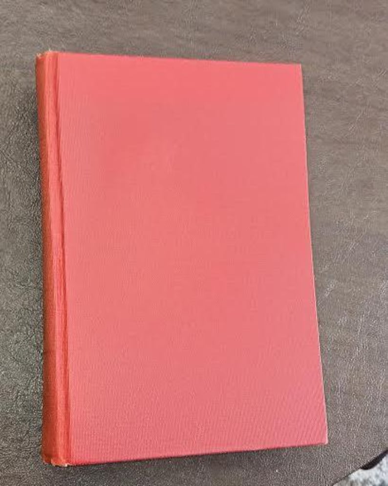 May include: A closed, hardback book with a solid coral-coloured cover. The spine is visible on the left side, and the pages are a similar shade. The book is rectangular and sits on a textured surface.