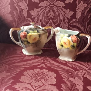 May include: A vintage floral tea set with a sugar bowl and creamer. The set is decorated with hand-painted roses and leaves in shades of yellow, pink, and green.