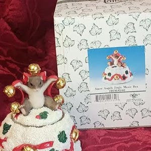 May include: A decorative music box featuring a mouse figurine with outstretched arms, adorned with gold bells and red ribbons. The mouse sits atop a snow-covered base with green holly accents. A box with a leaf pattern is in the background.