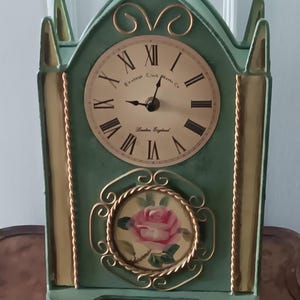 May include: A decorative green clock with gold accents. The clock face has Roman numerals and the text "Essence Clock Works Co. London, England." A rose painting is featured below the clock face.