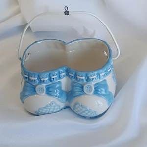 Puede incluir: Una maceta de cerámica con forma de patucos de bebé, pintada en blanco y azul claro. Los patucos tienen lazos azules, detalles de botones y un estampado de cuadros vichy en la puntera. Un asa de metal blanco está sujeta en la parte superior.