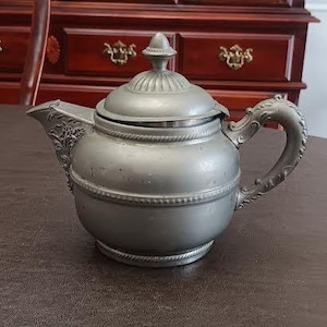 Op de afbeelding: Een zilverkleurige theepot met een deksel en een sierlijke handgreep. De theepot heeft een tuit en is versierd met gedetailleerde patronen. De handgreep heeft een gebogen ontwerp. De theepot staat op een bruin oppervlak.
