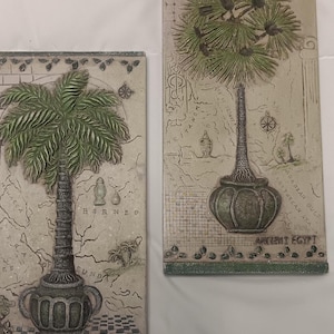 May include: Two rectangular wall art pieces with palm tree designs. The trees have green fronds and textured trunks, set against a beige background with a map design. The bases of the trees are a dark green colour. The words "ANCIENT EGYPT" are visible.