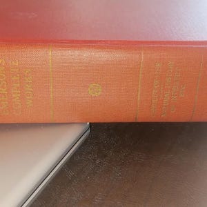 May include: A vintage book with an orange cover, featuring gold lettering that reads "Emerson's Complete Works" and "The Conduct of Life and Natural History". The book is resting on a light-coloured surface.