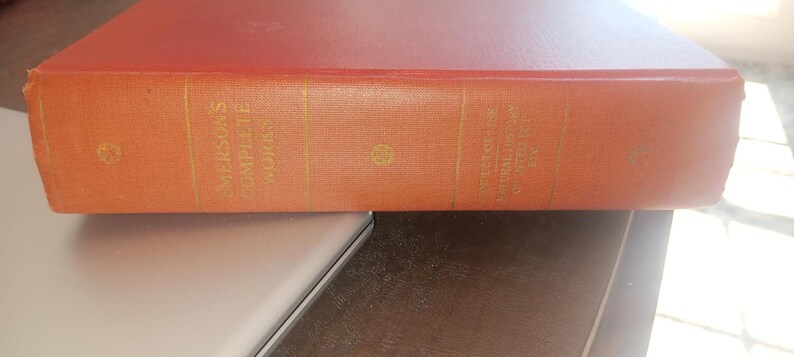 May include: A vintage book with an orange cover, featuring gold lettering that reads "Emerson's Complete Works" and "The Conduct of Life and Natural History". The book is resting on a light-coloured surface.