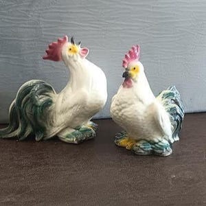 May include: Two ceramic rooster figurines. The roosters are white with pink combs and yellow accents. One rooster has a long, flowing tail with green and blue details. The other rooster has a shorter tail with similar coloring. Both are perched on a brown surface.