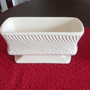 May include: A rectangular off-white ceramic planter featuring a raised diagonal wave pattern on the exterior sides. The vessel has a wide opening, a flared rim, and a solid base with a small beaded texture detail along the top and bottom edges.