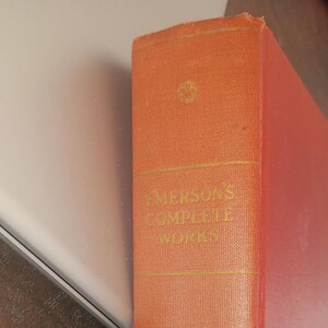 May include: Close-up of a vintage book spine. The book is bound in textured orange fabric with gold lettering. The title reads "Emerson's Complete Works". The book's edges are slightly worn, suggesting age.