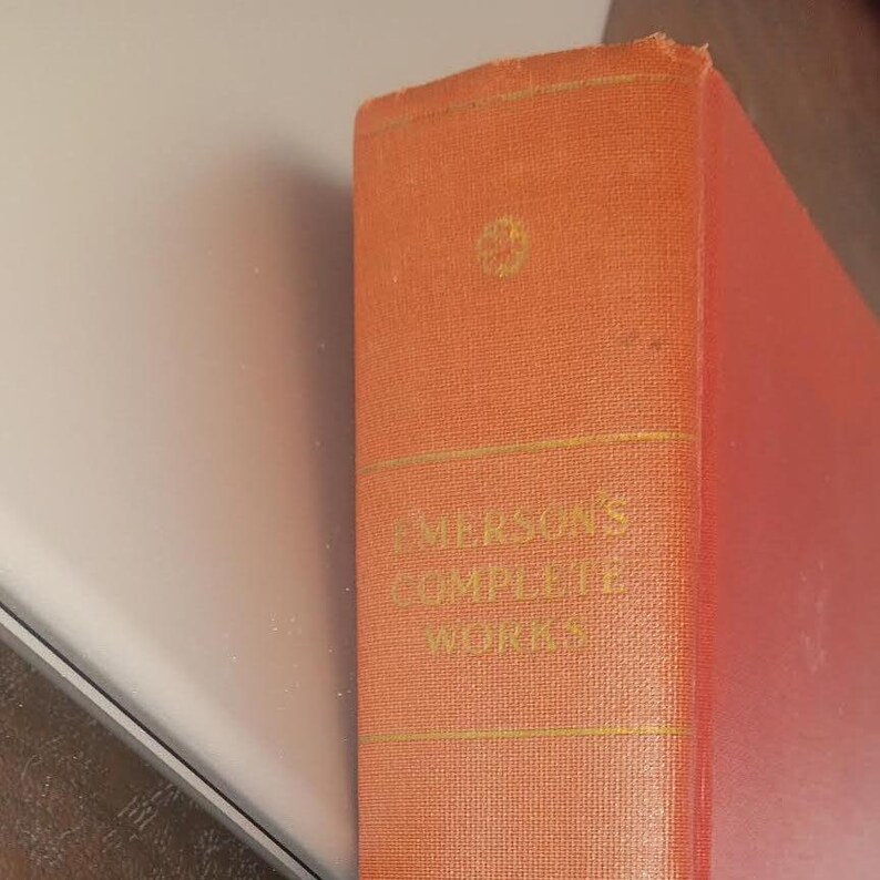 May include: Close-up of a vintage book spine. The book is bound in textured orange fabric with gold lettering. The title reads "Emerson's Complete Works". The book's edges are slightly worn, suggesting age.