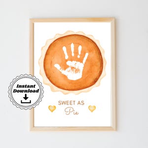Puede incluir: Una impresión de arte imprimible con una ilustración de acuarela de una tarta de calabaza con una huella de mano blanca en el centro. El texto "Sweet as Pie" está debajo de la tarta. Dos pequeños corazones amarillos están a cada lado del texto.