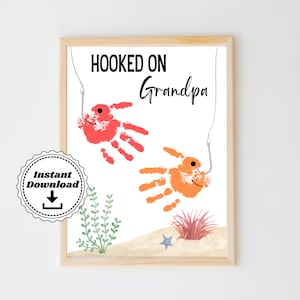 May include: A printable art print with the text "Hooked on Grandpa" and two handprint fish with fishing hooks. The fish are red and orange and are on a white background with a sandy beach and seaweed.