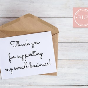 May include: A white card with the text "Thank you for supporting my small business!" in black cursive writing. The card is in a brown envelope on a white wooden background. A pink circle with the text "Beach Living Prints BLP Since 2022" is in the top right corner of the image.