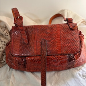 Può includere: Una borsa a mano rossa in pelle di serpente con chiusura a patta e una lunga tracolla regolabile. La borsa presenta una chiusura con fibbia e un manico sulla parte superiore. Il motivo a pelle di serpente è visibile all'esterno.