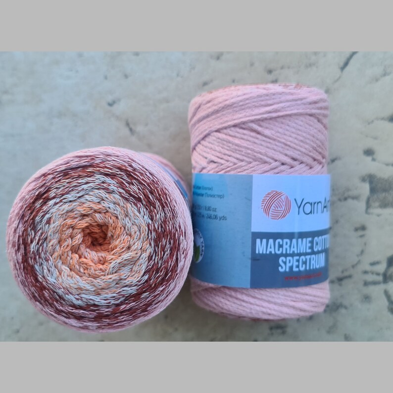 Puede incluir: Dos carretes de cuerda de macram&eacute;, uno de color rosa y el otro en un patr&oacute;n degradado multicolor de melocot&oacute;n, blanco y marr&oacute;n. La etiqueta en el carrete rosa dice "Yarn Arts Macrame Cotton Spectrum".