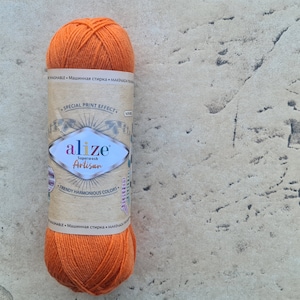 Può includere: Un gomitolo di filato arancione brillante con un'etichetta di carta. L'etichetta recita "alize Superwash Artisan" e "Trendy Harmonious Colors". Il filato è ben avvolto, adatto per progetti di maglia o uncinetto. Lo sfondo è una superficie strutturata e neutra.