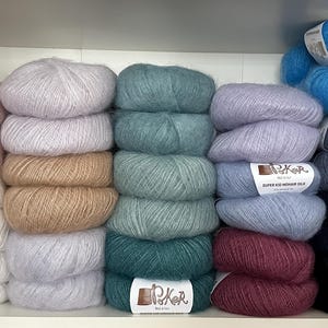 K&ouml;nnte beinhalten: Eine Sammlung flauschiger Garnkn&auml;uel in verschiedenen Farben, darunter Rosa, Wei&szlig;, Beige, Hellgrau, Blaugr&uuml;n, Blau, Lavendel, Burgunderrot, Dunkelgrau und Schwarz. Jedes Kn&auml;uel hat ein wei&szlig;es Papieretikett mit dem Text Poker Super Kid Mohair Silk.