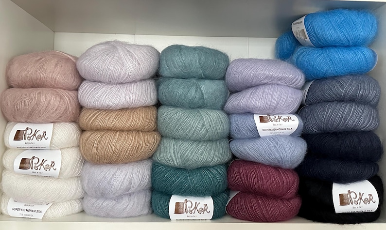 K&ouml;nnte beinhalten: Eine Sammlung flauschiger Garnkn&auml;uel in verschiedenen Farben, darunter Rosa, Wei&szlig;, Beige, Hellgrau, Blaugr&uuml;n, Blau, Lavendel, Burgunderrot, Dunkelgrau und Schwarz. Jedes Kn&auml;uel hat ein wei&szlig;es Papieretikett mit dem Text Poker Super Kid Mohair Silk.